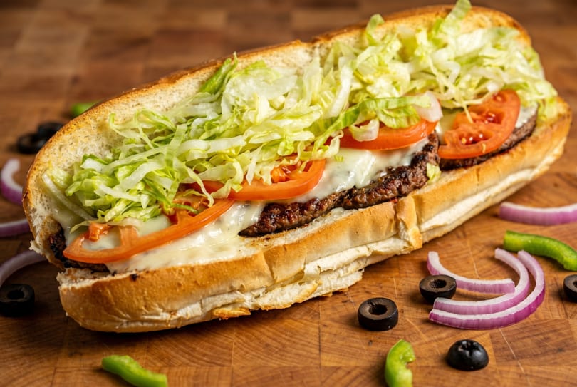 A large sub sandwich filled with meat, melted cheese, tomatoes, and shredded lettuce on a wooden board.