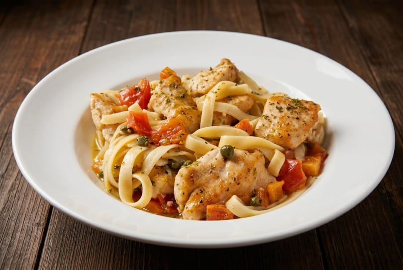Chicken pasta with tomatoes and capers in a white bowl on a rustic wooden table.