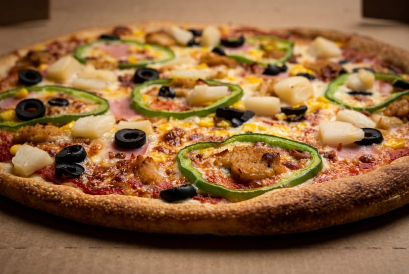 Close-up of a pizza with green peppers, black olives, pineapple, and chicken on a brown surface.
