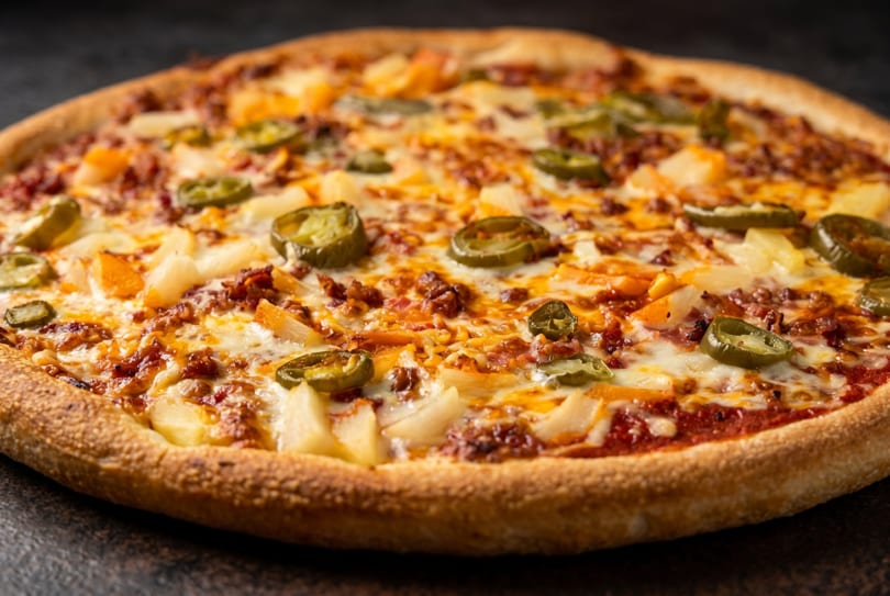Close-up of a pizza with melted cheese, pineapple, ground meat, and jalapeño slices.